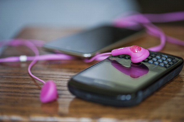 A Blackberry cell phone, ipod and pink earbuds lie on a wood table.